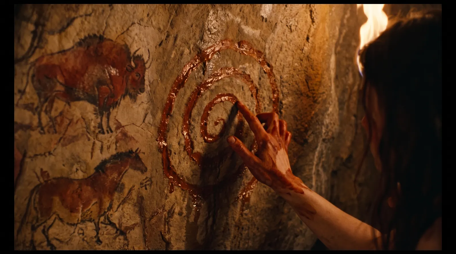 A hand smeared with ochre traces a first spiral on a cave wall